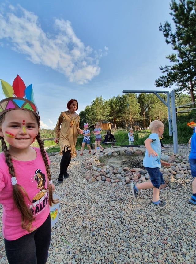 Dziewczynka z pomalowaną twarzą i pióropuszem, w tle dzieci i kobieta przebrana za Indianina, zabawa tematyczna na dworze.