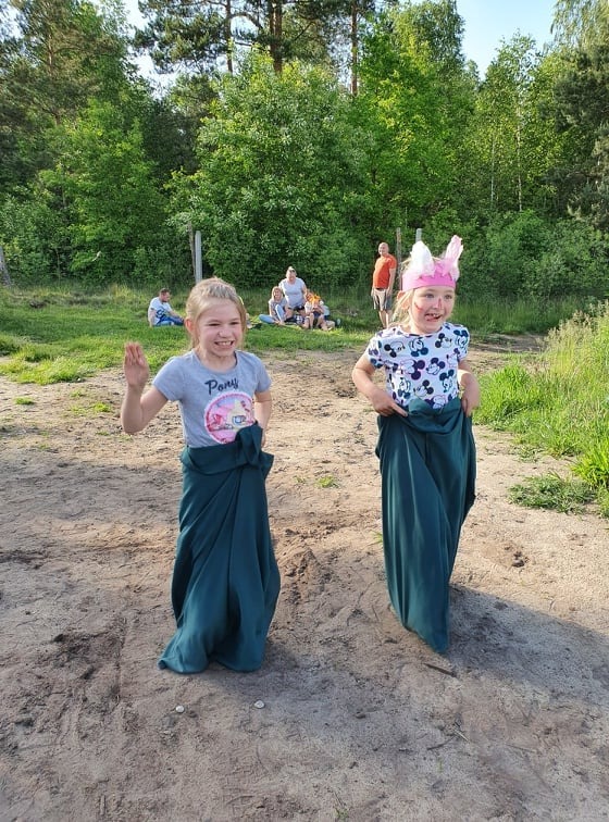 Dwie uśmiechnięte dziewczynki w workach do skakania, jedna macha ręką, druga ma opaskę z uszami królika, w tle trawa, drzewa i grupa osób.