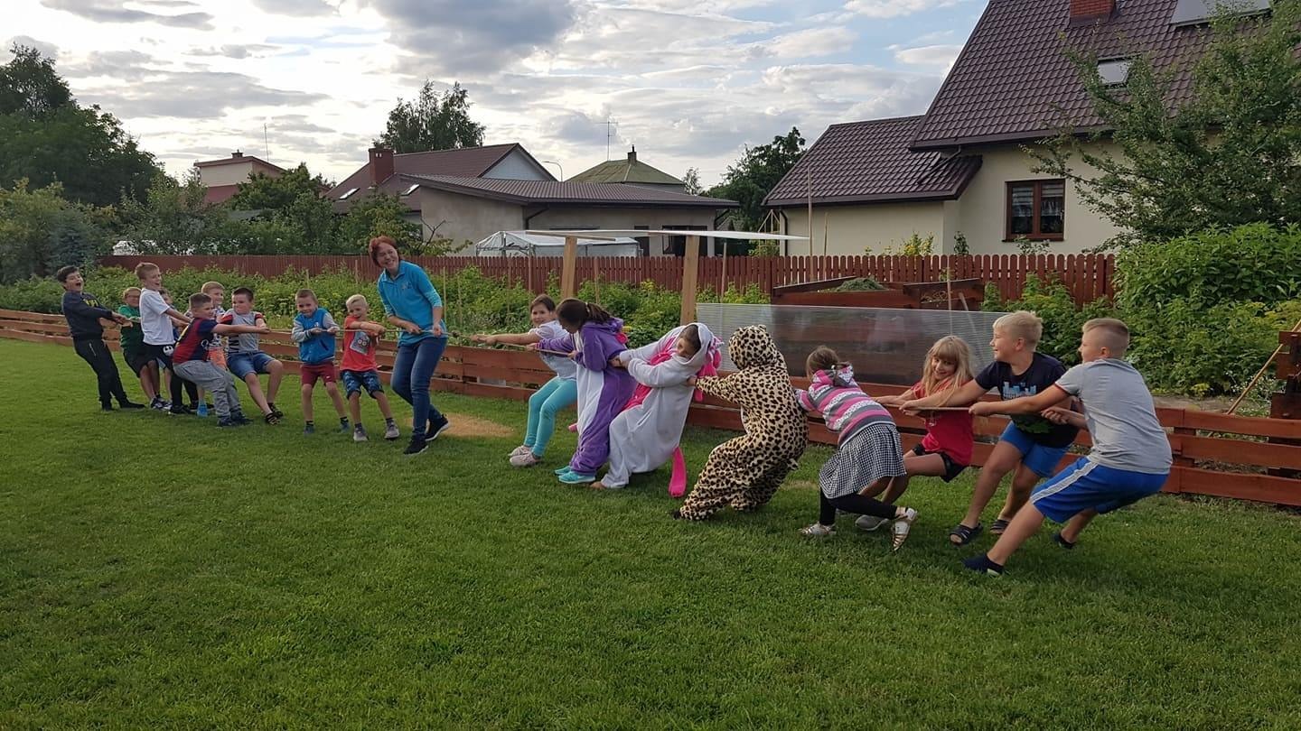 Grupa dzieci w różnym wieku, niektóre w kostiumach (jednorożec, panterka), bierze udział w przeciąganiu liny na trawniku przed domem jednorodzinnym w słoneczny dzień.