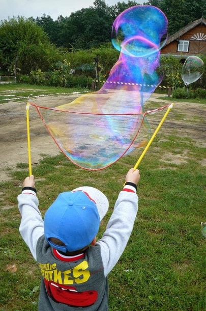 Chłopiec w Warszawie, ubrany w bluzę z motywem Cars, puszcza ogromne bańki mydlane za pomocą specjalnego sznurka i kijków. W tle widoczny dom i drzewa.
