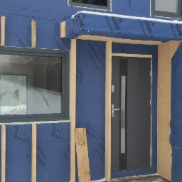 Montaż okna PCV i drzwi wejściowych w nowym domu w trakcie budowy, widoczne ocieplenie ścian folią paroizolacyjną i drewniane ramy okienne.