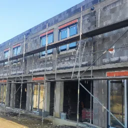 Budynek w trakcie budowy, z zamontowanymi oknami z roletami zewnętrznymi, widoczne rusztowania i materiały budowlane.