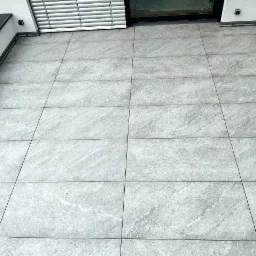 Wykończony balkon z szarymi, kwadratowymi płytkami imitującymi kamień, ułożonymi równo z ciemnymi fugami, widok z góry.