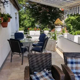 Umeblowany taras z markizą, szklanym stolikiem i rattanowymi fotelami z poduszkami w kratę, donice z kwiatami w tle.