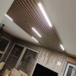 Drewniana zabudowa sufitu z oświetleniem LED, częściowo widoczna ściana z dekoracyjnymi drewnianymi listwami, okno z żaluzjami i telewizor na ścianie. W tle widoczne kartony.