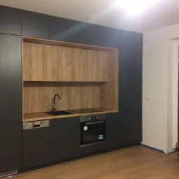 Nowoczesna, minimalistyczna kuchnia z ciemnoszarymi szafkami, drewnianymi akcentami, czarnym zlewem i płytą grzewczą, oświetleniem LED podszafkowym i choinką w tle.