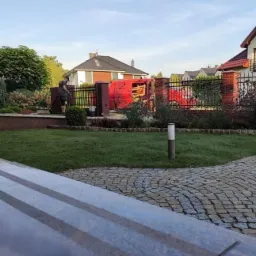 Świeżo założony trawnik w ogrodzie z kostki brukowej, z widocznym ogrodzeniem i pracownikiem w tle, obok zaparkowany czerwony samochód dostawczy.