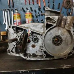 Otwarty, brudny blok silnika motocyklowego na warsztatowym stole, widoczne wewnętrzne elementy mechanizmu, narzędzia i preparaty w tle.