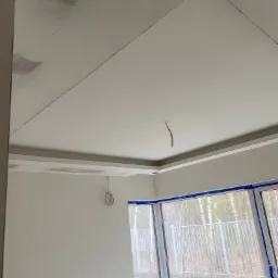 Niedokończony sufit podwieszany z płyt kartonowo-gipsowych, widoczne łączenia i ślady szpachlowania, okno zabezpieczone folią malarską, wystające przewody elektryczne.