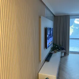 Ściana z dekoracyjnymi panelami pionowymi w odcieniach beżu, telewizor LCD zawieszony na szarej płycie, biała szafka RTV z rośliną doniczkową, parkiet ułożony w jodełkę, zasłony w tle.