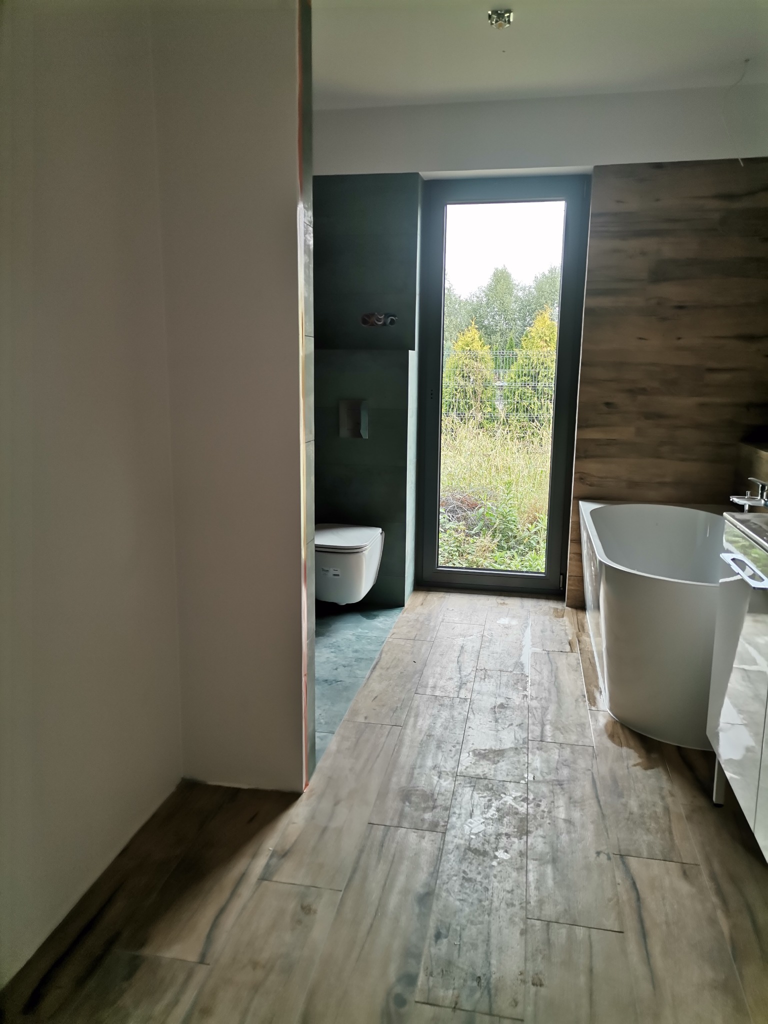 Nowoczesna łazienka z drewnopodobną podłogą, białą wanną i toaletą w zabudowie, widok na zieleń przez okno, ślady po pracach na podłodze.