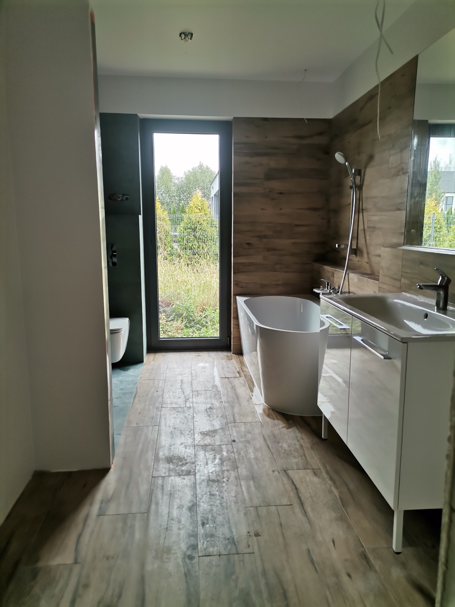 Nowoczesna łazienka z wolnostojącą białą wanną, umywalką na szafce w kolorze białym połysku, drewnopodobnymi płytkami na ścianie i podłodze, oraz oknem z widokiem na ogród.