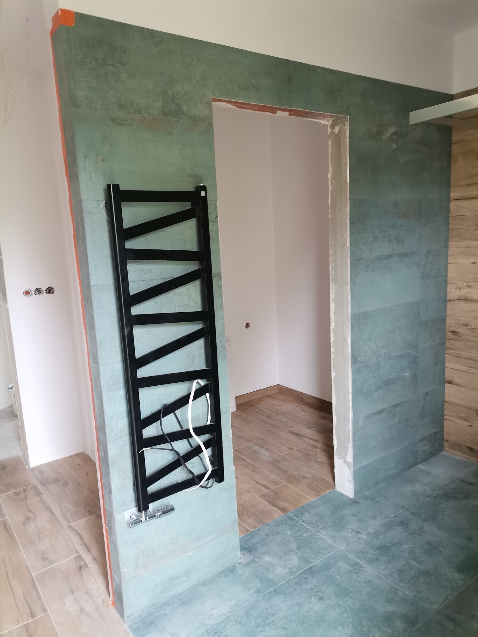 Łazienka w trakcie remontu: ściany wyłożone turkusowymi płytkami, zamontowany czarny grzejnik drabinkowy, widoczne niedokończone obramowanie drzwi i podłoga z paneli.