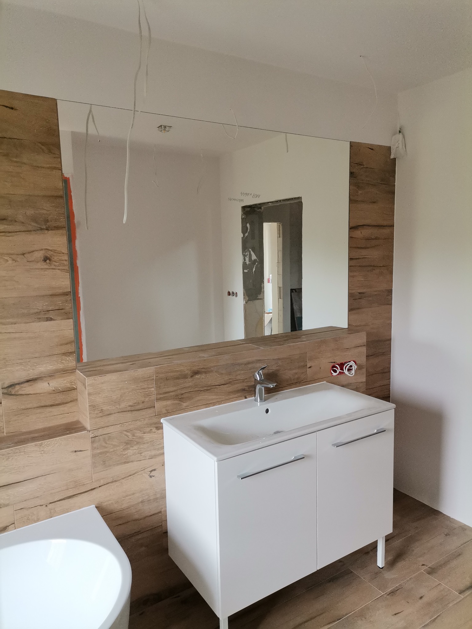 Łazienka w trakcie remontu: umywalka z szafką, lustro z drewnopodobnymi płytkami, widoczne kable wystające z sufitu i niezakończone prace wykończeniowe w przejściu do innego pomieszczenia.