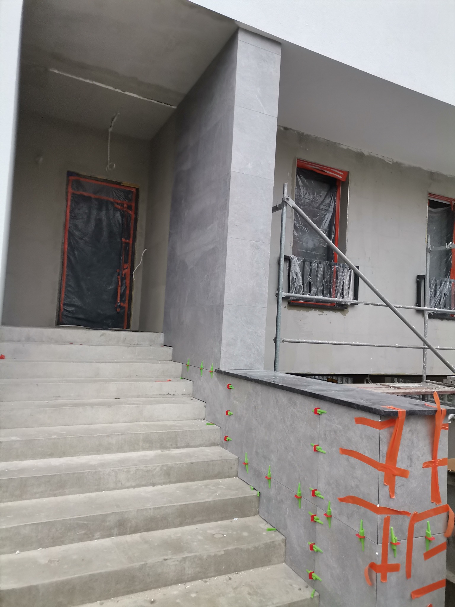 Montaż szarych płytek elewacyjnych na schodach wejściowych do budynku, widoczne poziomice i plastikowe krzyżyki dystansowe.