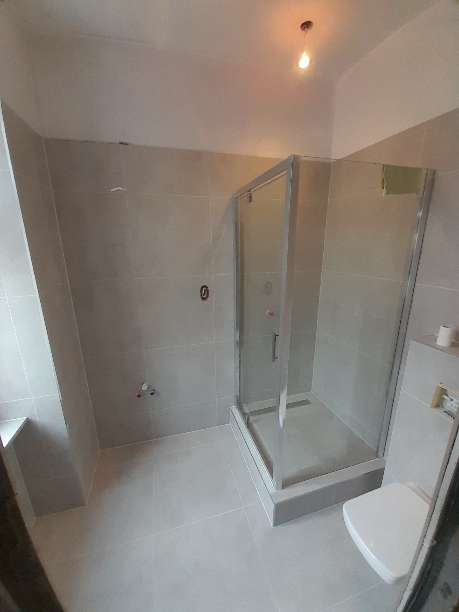 Jasna łazienka po remoncie z kabiną prysznicową z szarą ramą, widoczne płytki w odcieniach szarości, biała toaleta i fragment okna z parapetem.