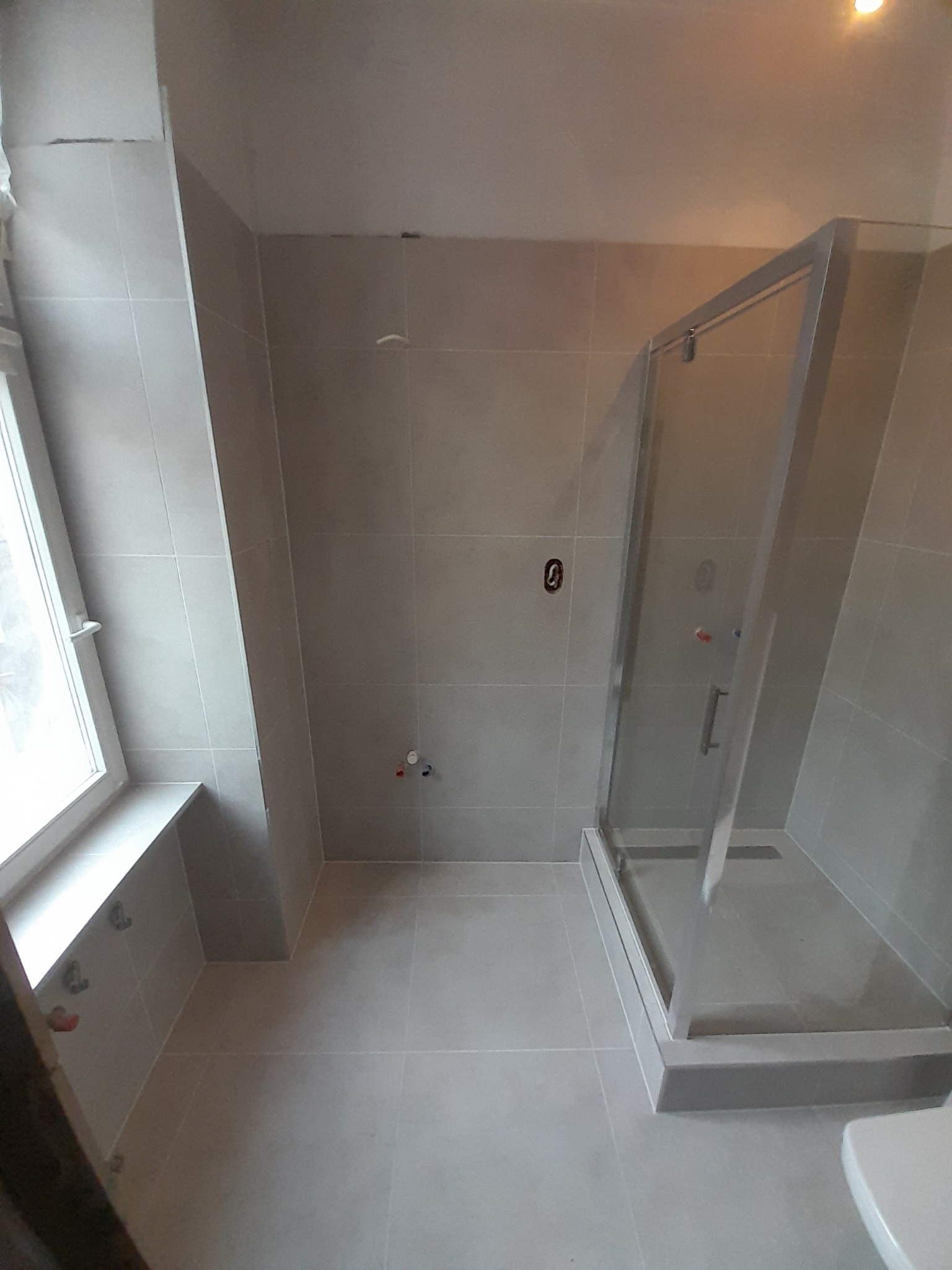 Wykończona łazienka w szarych płytkach z kabiną prysznicową, widoczne przyłącza wodne i okno z parapetem.