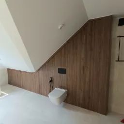 Łazienka na poddaszu z zabudową z paneli drewnopodobnych, białą toaletą podwieszaną i wanną z podświetleniem LED u dołu.