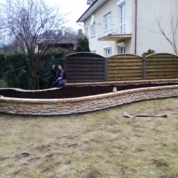 Mark.Kom Rafał Klementowski - Mur oporowy z piaskowca, wypełniony ziemią, z siedzącym na nim mężczyzną i łopatą na trawie, na tle domu i ogrodzenia.