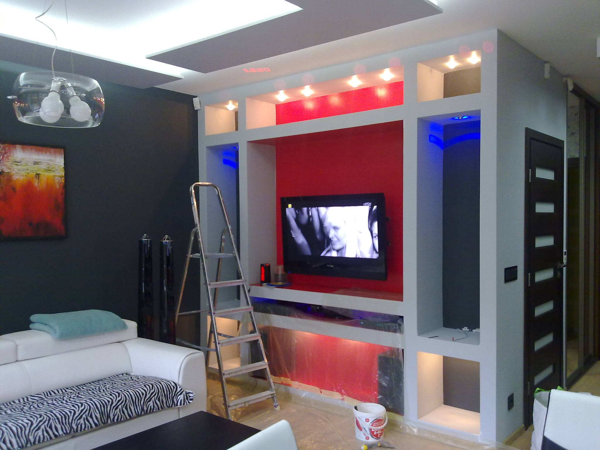 Remontowany salon w Markach: ściana pod telewizor z oświetleniem LED w trakcie malowania, widoczna drabina malarska, folia ochronna, pędzel i wiadro z farbą.