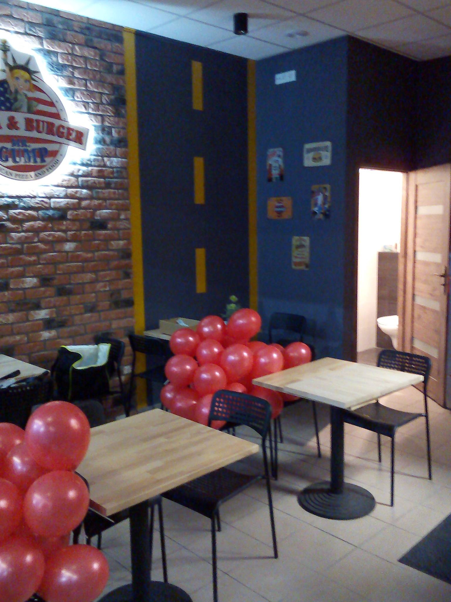 Wnętrze lokalu gastronomicznego z dekoracyjną ścianą z cegły, niebieską ścianą z żółtymi pasami, drewnianymi stolikami, czarnymi krzesłami i czerwonymi balonami, widoczny fragment toalety.