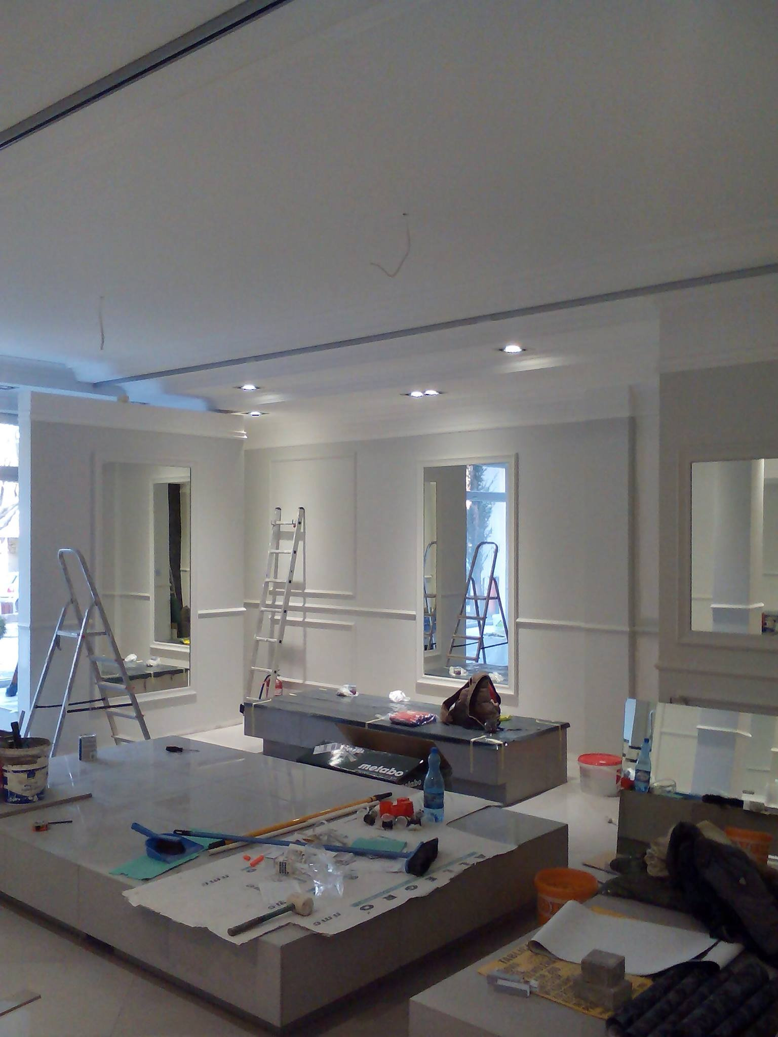 Wnętrze w trakcie prac wykończeniowych: białe ściany z listwami dekoracyjnymi, lustra w ozdobnych ramach, rozstawione drabiny, powierzchnie zabezpieczone folią, widoczne narzędzia i materiały...