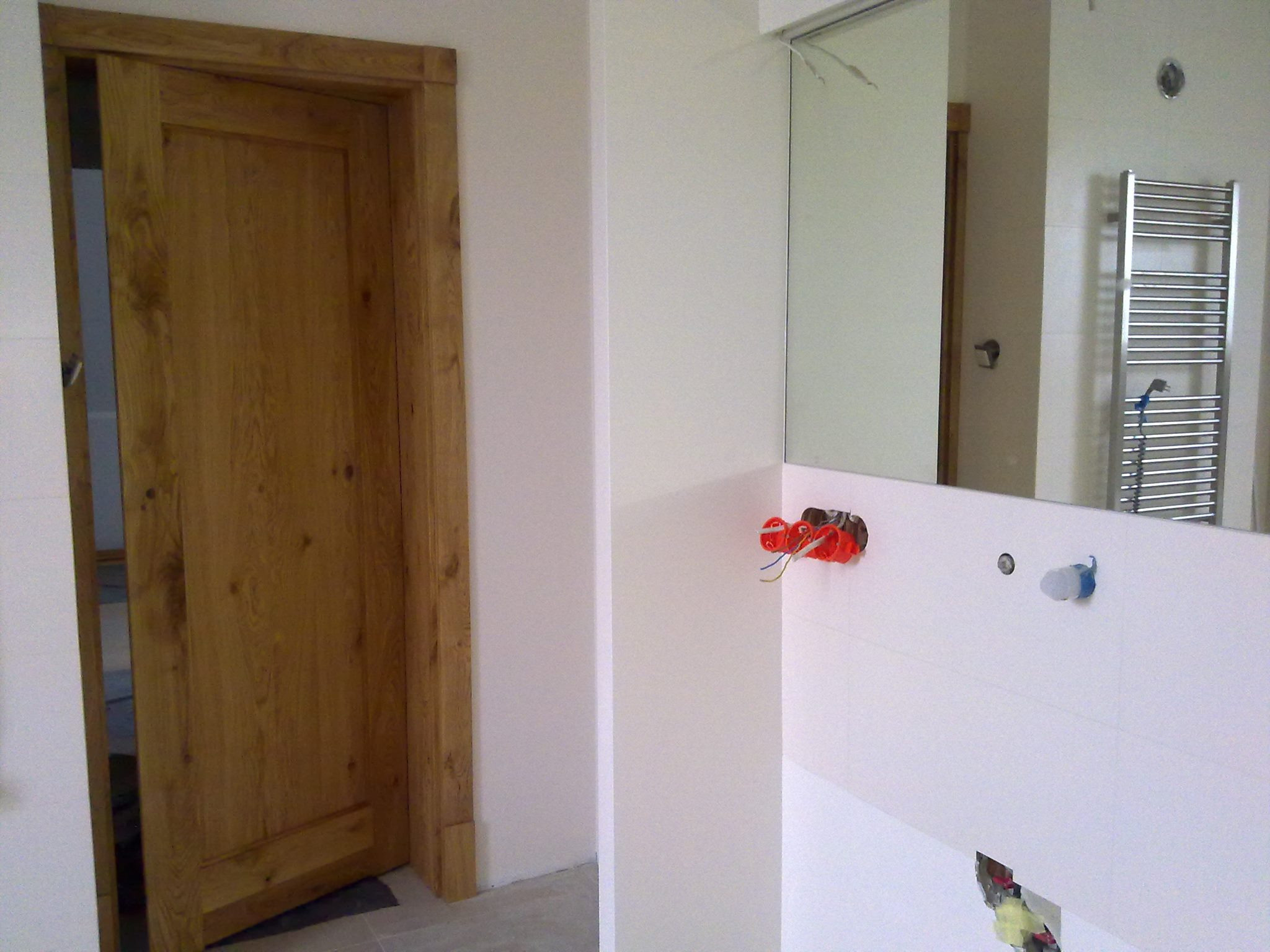 Wnętrze łazienki w trakcie remontu: widoczna drewniana ościeżnica z drzwiami, białe płytki na ścianach, lustro oraz wystające kable elektryczne i rury hydrauliczne.