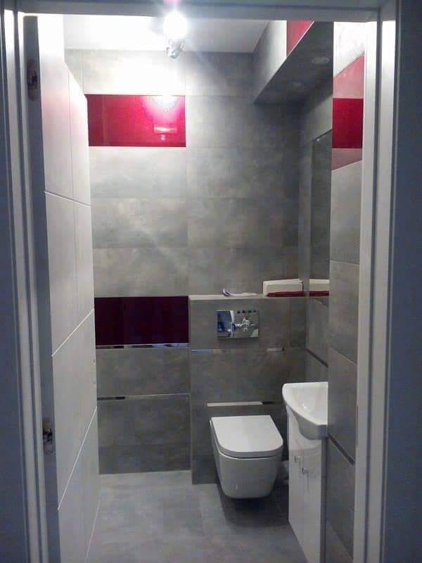 Nowoczesna łazienka z szarymi płytkami imitującymi beton, akcentami z czerwonego szkła, podwieszaną toaletą i umywalką z szafką, widziana zza futryny drzwi.