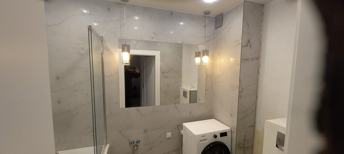 Wykończona łazienka z marmurowymi płytkami, szklanym panelem prysznicowym, wiszącymi lampami i pralką, widoczne przyłącza wody.