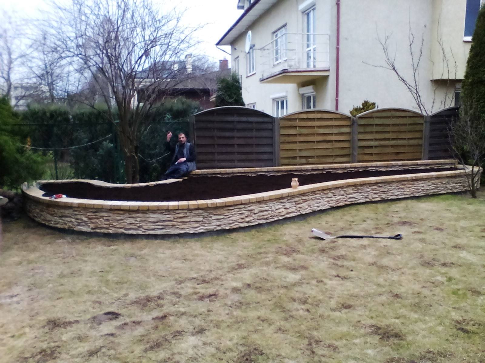 Mur oporowy z piaskowca, wypełniony ziemią, z siedzącym na nim mężczyzną i łopatą na trawie, na tle domu i ogrodzenia.