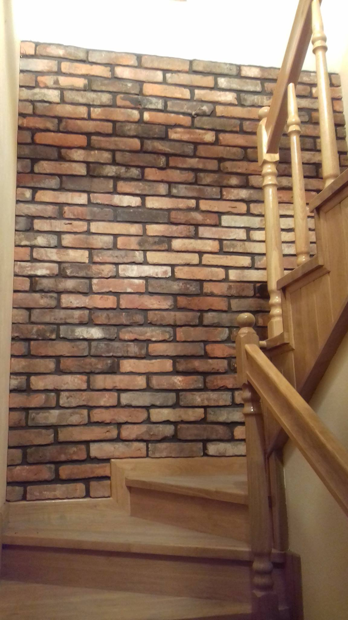 Ściana wyłożona dekoracyjną cegłą w odcieniach czerwieni i brązu, sąsiadująca z drewnianymi schodami z balustradą