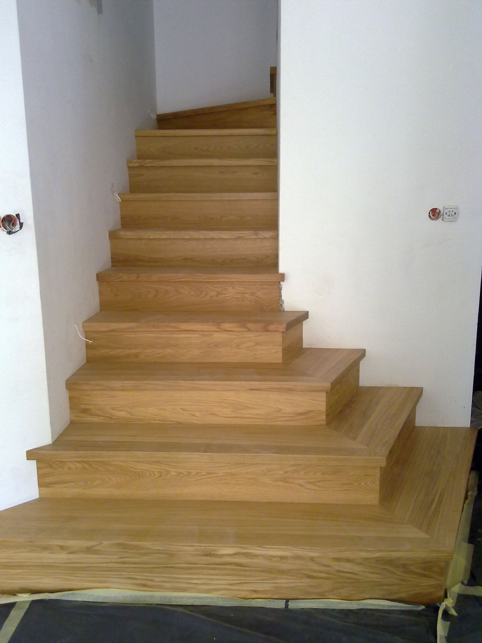 Drewniane schody dębowe z widocznym usłojeniem, montowane przy białej ścianie z wystającymi przewodami elektrycznymi i zabezpieczoną podłogą.