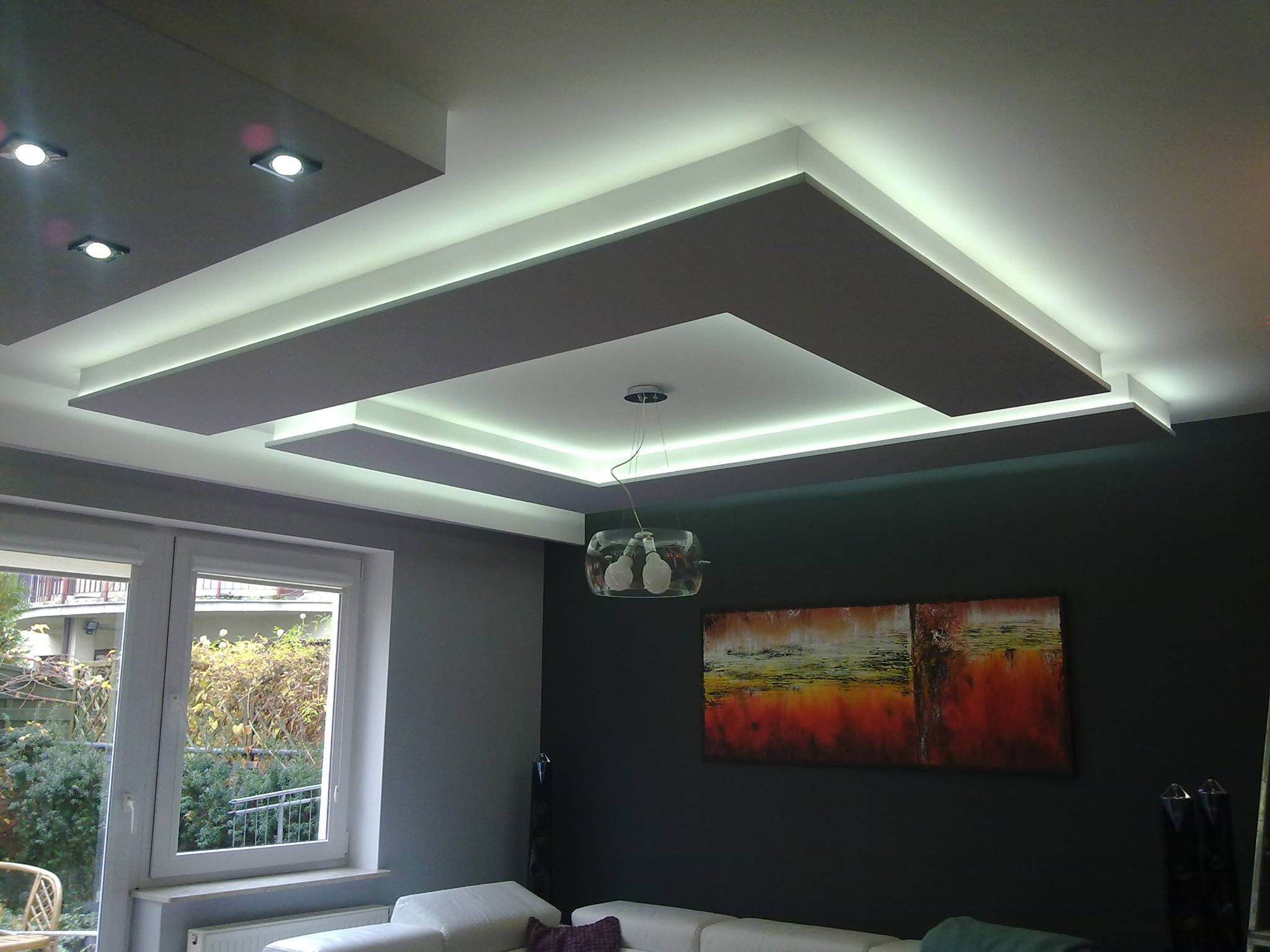 Nowoczesny salon z podwieszanym sufitem z oświetleniem LED, szare ściany, okno z widokiem na ogród, abstrakcyjny obraz na ścianie.