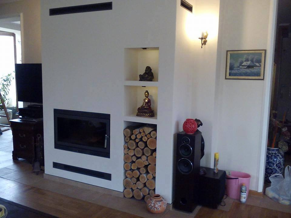 Nowoczesny salon z kominkiem i dekoracyjnymi niszami, w których umieszczono drewno opałowe i figurki Buddy. Na ścianie kinkiet i obraz. Podłoga wyłożona drewnianymi panelami.