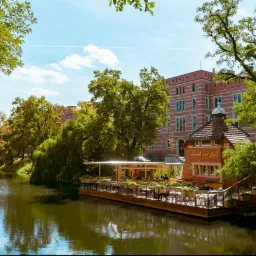 Restauracja na wodzie z drewnianym tarasem, otoczona zielenią i ceglanym budynkiem w tle.