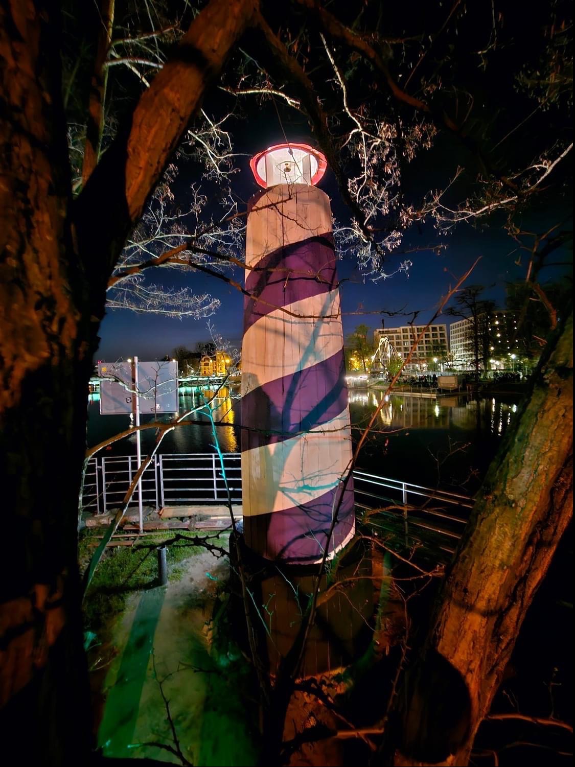 Instalacja artystyczna: Latarnia morska w biało-fioletowe pasy, oświetlona od góry, widziana nocą przez gałęzie drzew, odbijająca się w wodzie stawu, w tle budynki i oświetlony diabelski młyn.