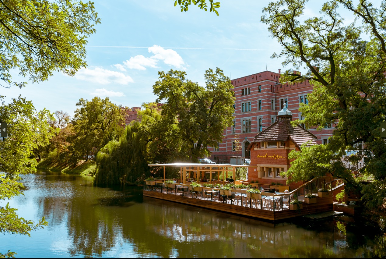 Restauracja na wodzie z drewnianym tarasem, otoczona zielenią i ceglanym budynkiem w tle.