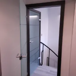 Szklane drzwi z poziomymi pasami w ciemnej ramie, prowadzące na schody z czarną balustradą, widoczne z perspektywy wnętrza.