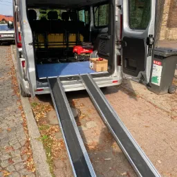 Srebrny van z otwartymi tylnymi drzwiami i wyciągniętymi rampami dla wózków inwalidzkich, widoczne siedzenia i przedmioty wewnątrz, na zewnątrz kostka brukowa i liście.