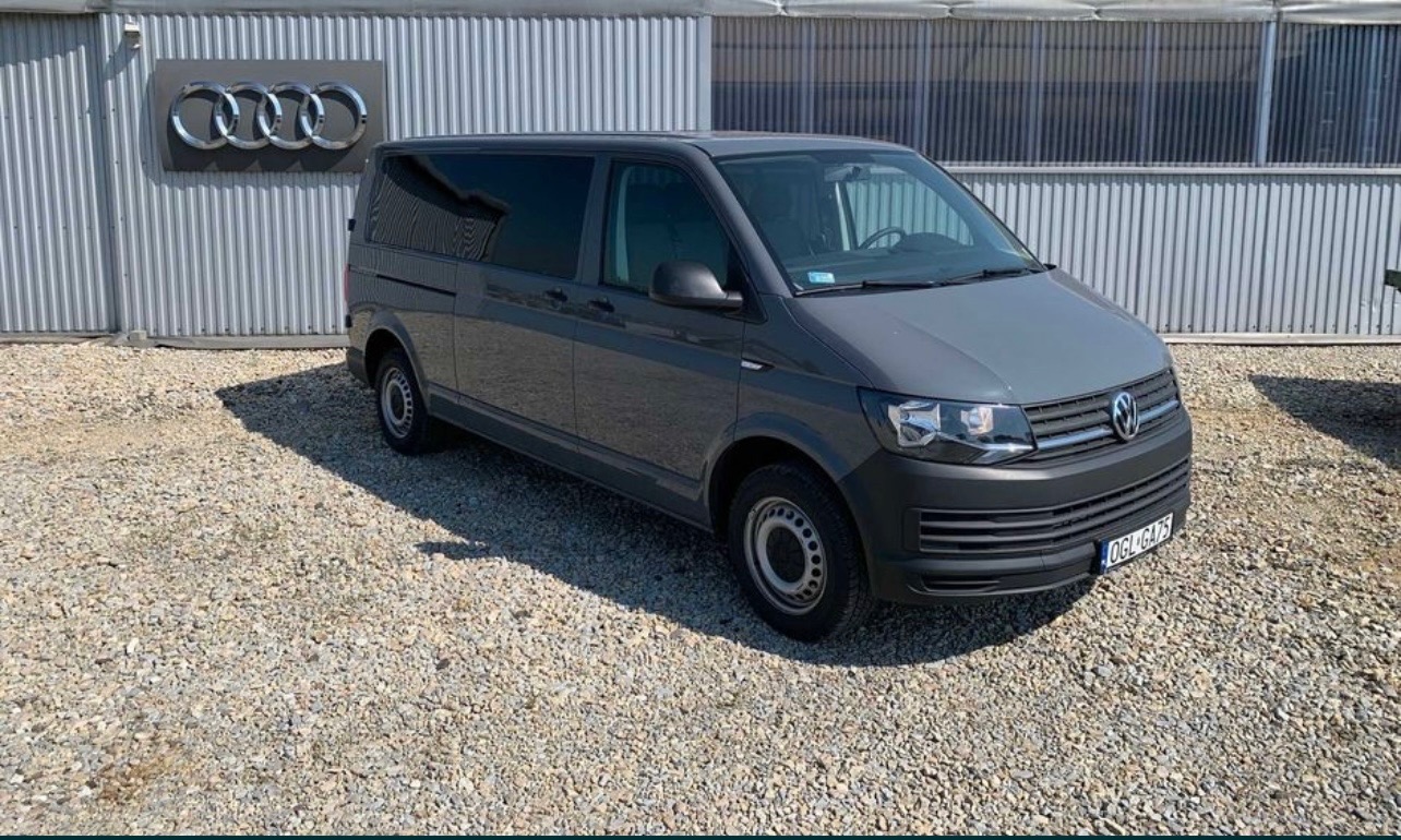 Szary Volkswagen Transporter T6 z przyciemnianymi szybami, zaparkowany na żwirowym placu przed szarym budynkiem z logo Audi na ścianie.