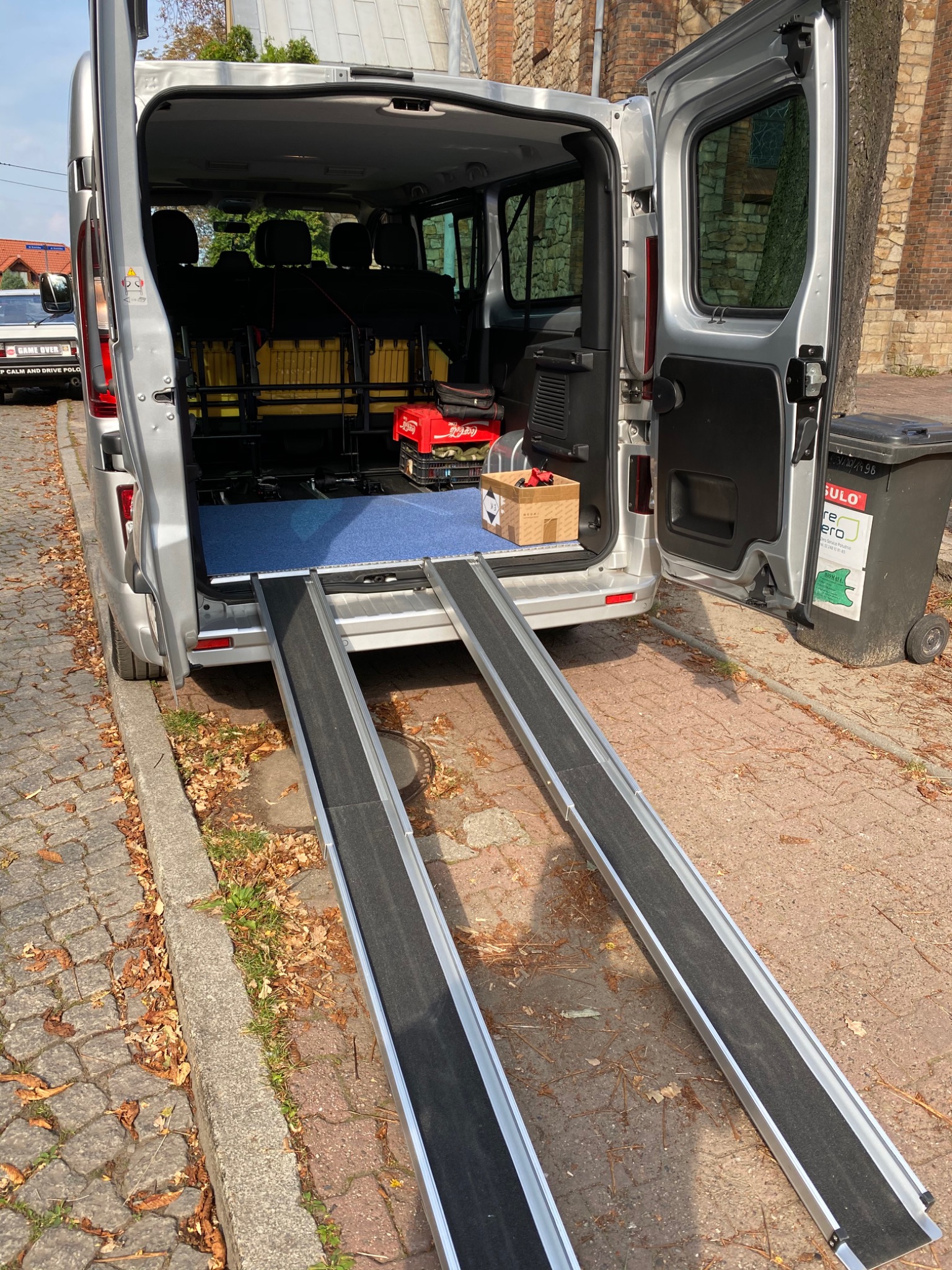 Srebrny van z otwartymi tylnymi drzwiami i wyciągniętymi rampami dla wózków inwalidzkich, widoczne siedzenia i przedmioty wewnątrz, na zewnątrz kostka brukowa i liście.