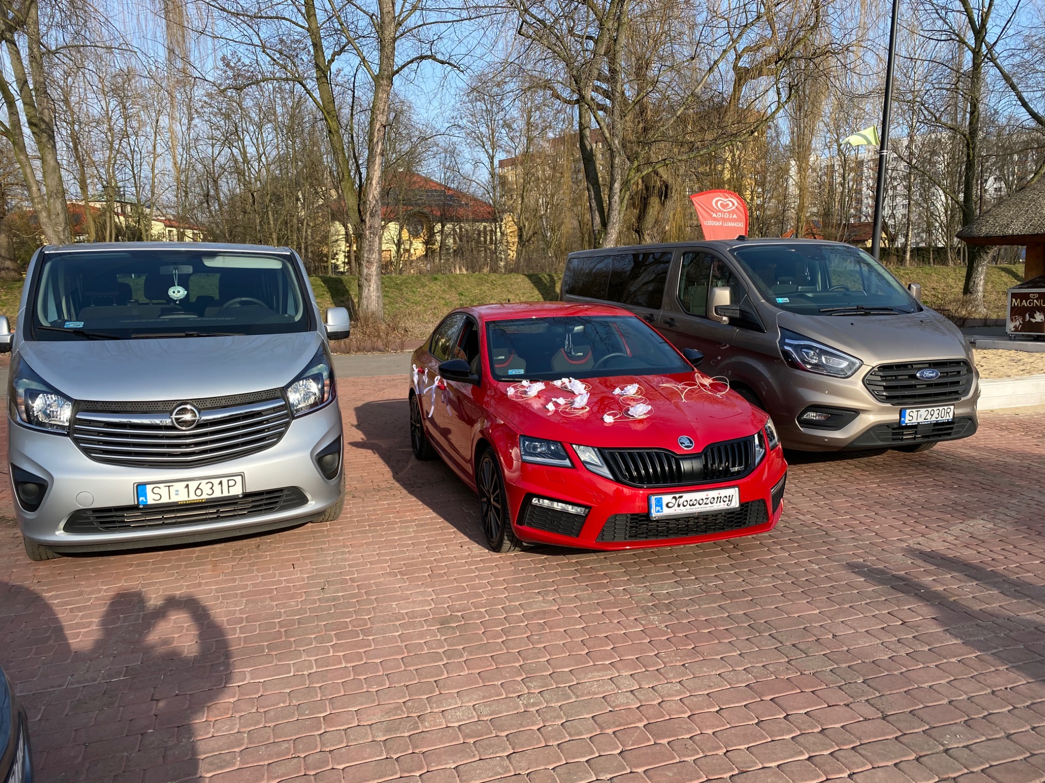 Trzy samochody: srebrny van Opel Vivaro, czerwona Skoda Octavia z dekoracjami ślubnymi na masce i brązowy Ford Transit Custom, stojące na brukowanym parkingu w słoneczny dzień.