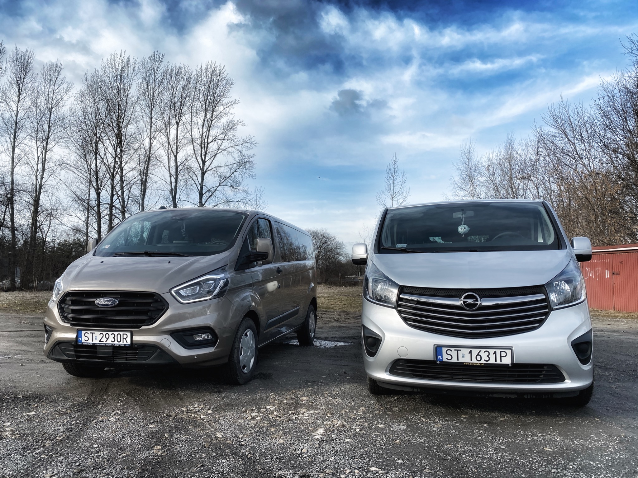 Dwa busy: beżowy Ford Transit Custom z numerem rejestracyjnym ST 2930R i srebrny Opel Vivaro z numerem rejestracyjnym ST 1631P, zaparkowane na żwirowym placu w pochmurny dzień.