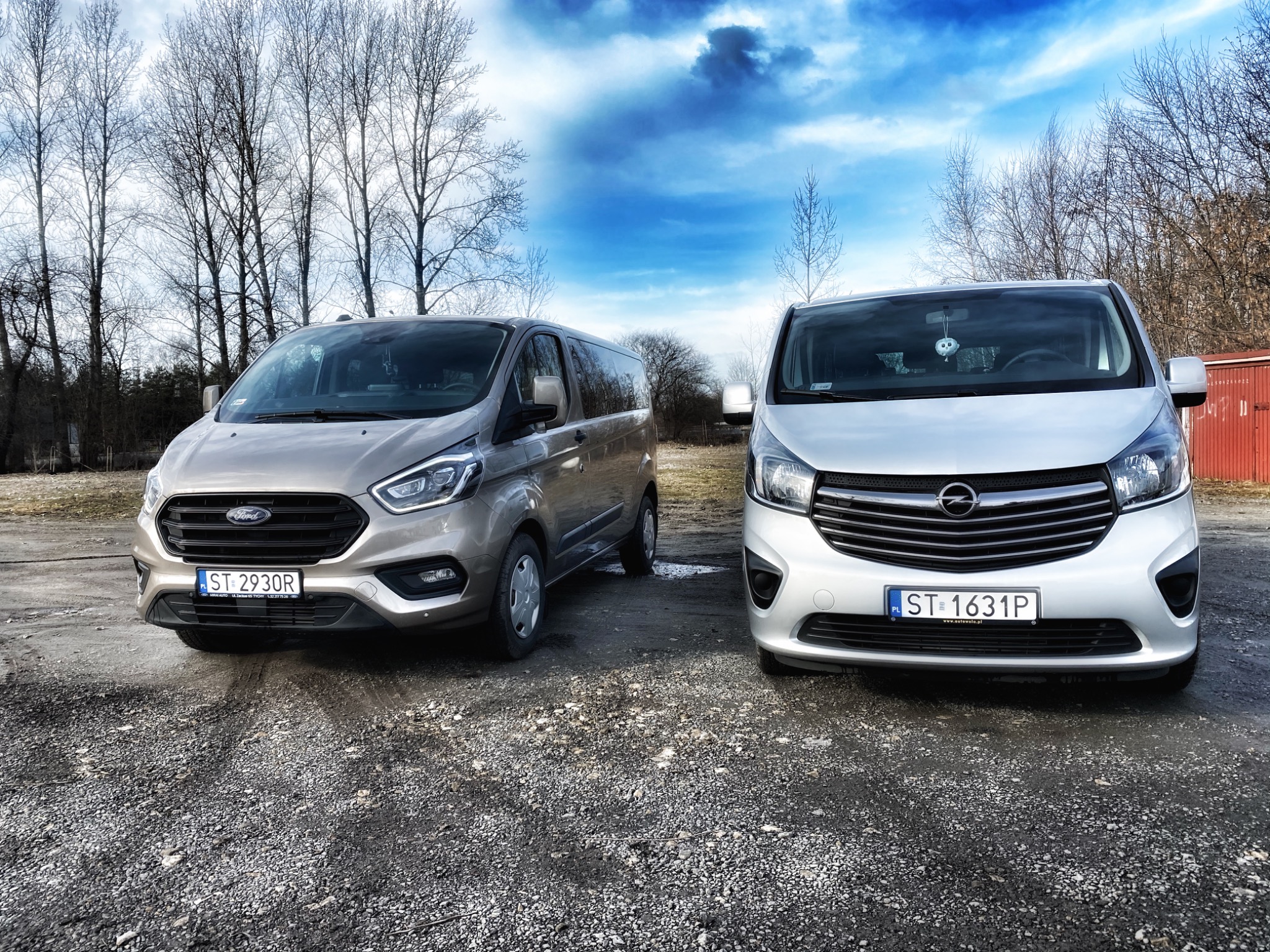 Dwa busy: beżowy Ford Transit Custom i srebrny Opel Vivaro, zaparkowane na żwirowym placu, widoczne z przodu, z polskimi tablicami rejestracyjnymi i drzewami w tle pod pochmurnym niebem.