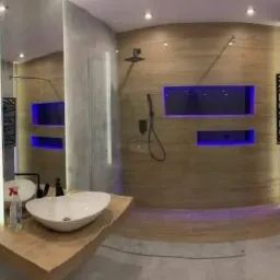 Nowoczesna łazienka z drewnianymi panelami, podświetlanymi wnękami w kolorze niebieskim, umywalką nablatową i grzejnikiem drabinkowym.