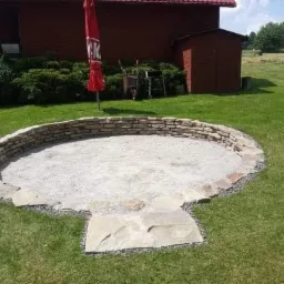 Okrągłe palenisko z kamienia na trawniku, częściowo wypełnione żwirem, z widoczną czerwoną altaną w tle.