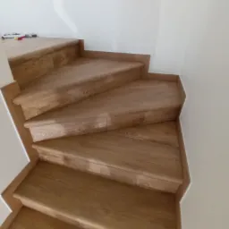 Drewniane schody z wykończeniem w kolorze jasnego dębu, widoczne z góry, z białymi ścianami w tle, częściowo wykończone.