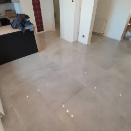 Salon z nową, jasną, błyszczącą podłogą z płytek ceramicznych w odcieniach szarości, widoczny fragment kuchni z czarną zabudową i drewnianym blatem.