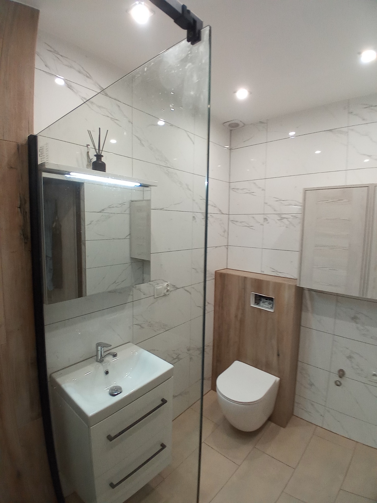 Nowoczesna łazienka z białymi marmurkowymi płytkami, wiszącą toaletą zabudowaną drewnianą płytą, szafką z umywalką i szklaną kabiną prysznicową, widoczny dyfuzor zapachowy na szafce z lustrem.