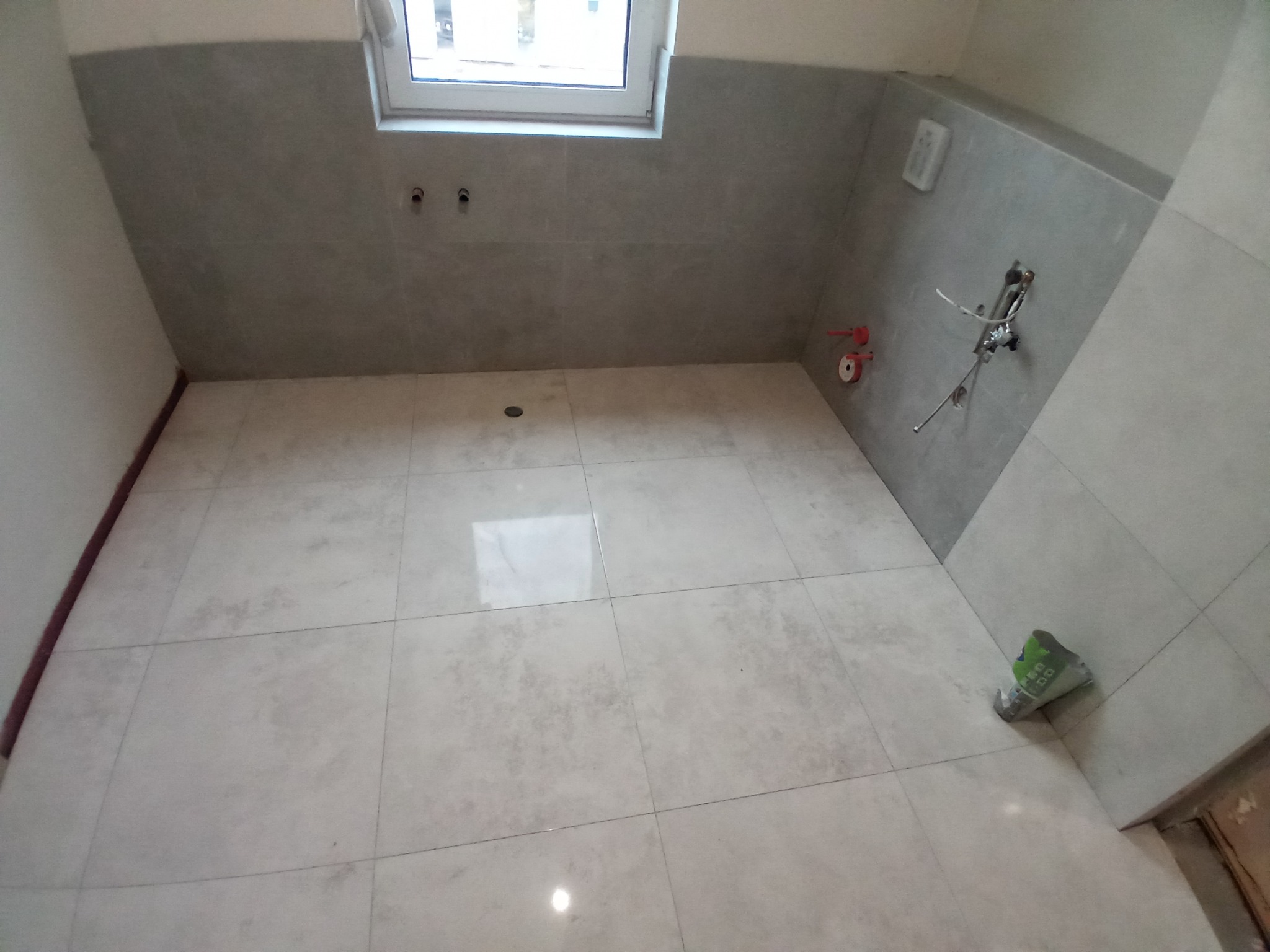 Świeżo wykafelkowana łazienka z szarymi płytkami na ścianach i podłodze, widoczne przygotowane instalacje wodne i kanalizacyjne, oraz resztki opakowania po zaprawie.